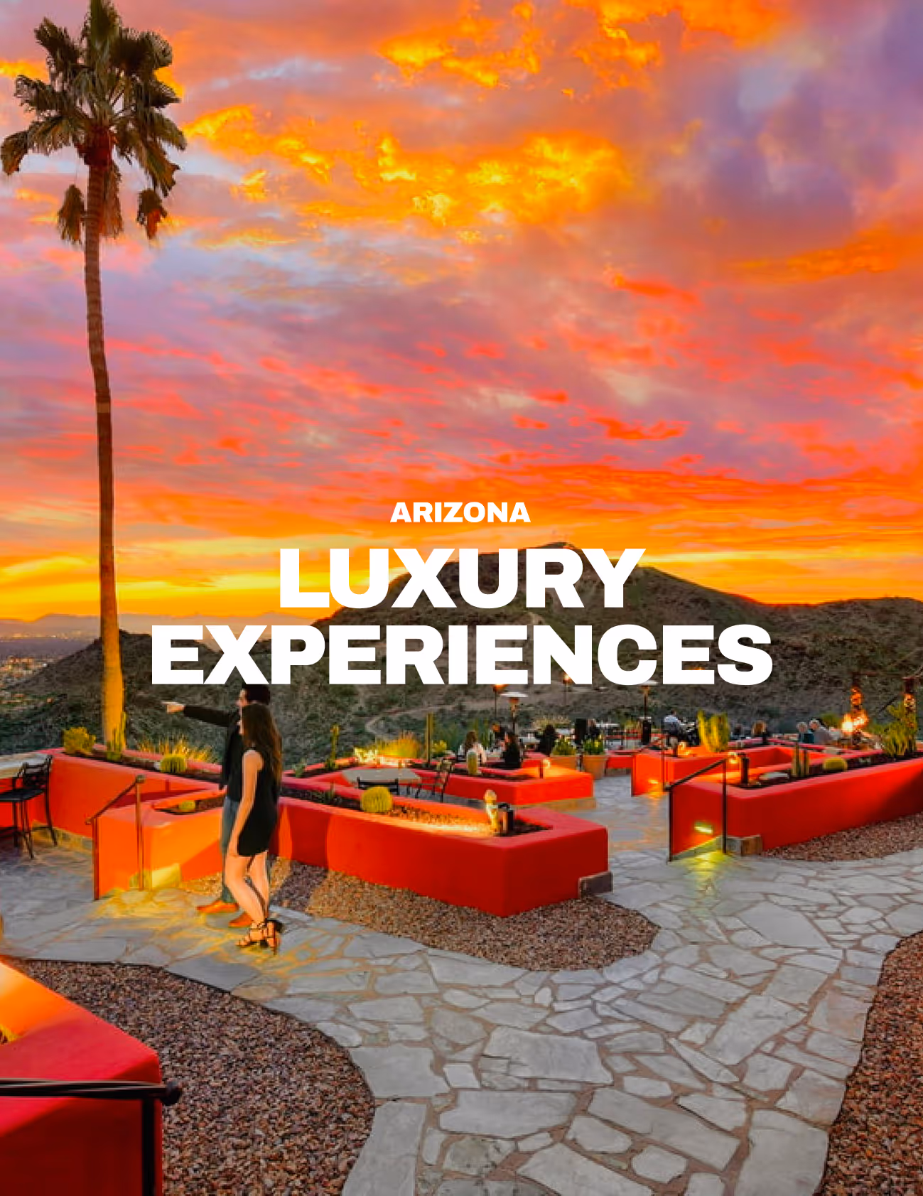 Couple walking on stone pathway through red outdoor lounge seating under a vibrant orange sunset sky with desert hills in Arizona.